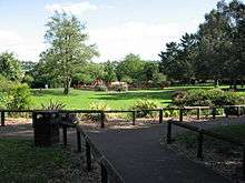 Area with grass and trees. In the distance is a children's play area and in the foreground a path with wooden rails.