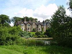 Country house, partially obscured by greenery, viewed from across a pond. The observable frontage comprises five gables, with a turret between the four gables on the left and the rightmost gable.