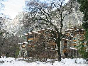 Photograph of The Ahwahnee in winter snow, amidst bare trees with the dramatic walls of Yosemite Valley rising behind.