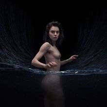 A nude Leanne Macomber stands with the bottom-half of her body in water.