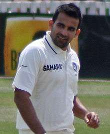 Indian fast-bowler Zaheer Khan walking up to his run-up in the middle of an over. Khan is a dark skinned person, wearing a white shirt. The cricket field can be seen in the background.