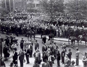 Several police officers on horses watch a large crowd standing in a public park.